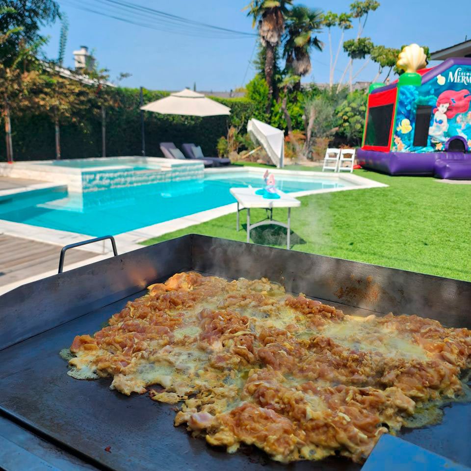 Home Tacolab LA live grill catering at a poolside party in Los Angeles. Fresh chicken sizzling on the flat top with inflatable bouncy castle and swimming pool in the background.